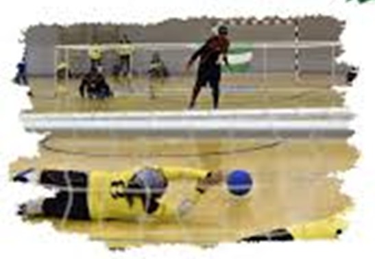 Escena de goalball en un pabellón, con varias personas frente a una portería en el fondo. En primer plano, una persona está tumbada en el suelo intentando bloquear una pelota azul.