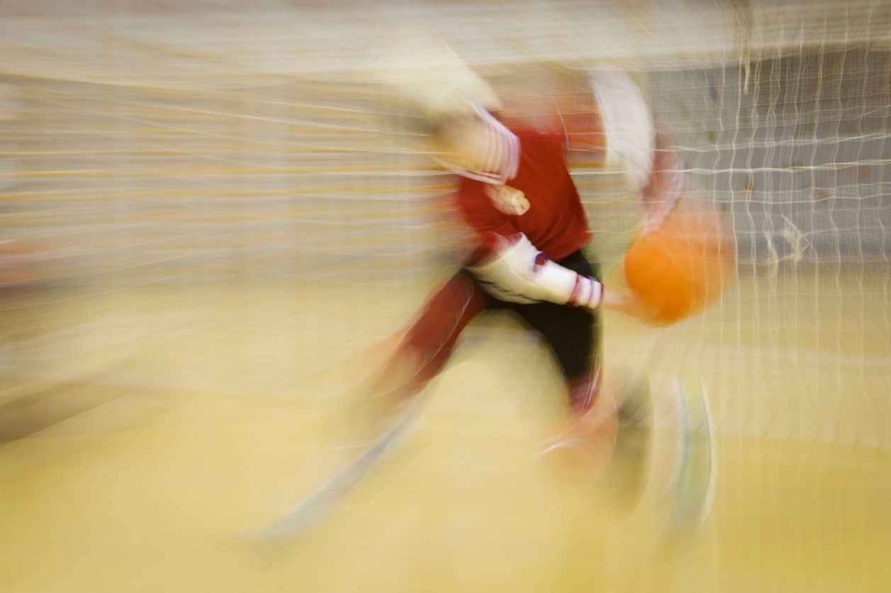 La imagen muestra a un jugador de goalball captado en pleno movimiento, con un efecto de desenfoque que transmite gran velocidad durante el lanzamiento. El balón naranja se distingue a la derecha mientras el cuerpo se inclina hacia delante, próximo a la portería. El fondo incluye la red y el suelo de pista, difuminados para enfatizar la acción dinámica del deportista.