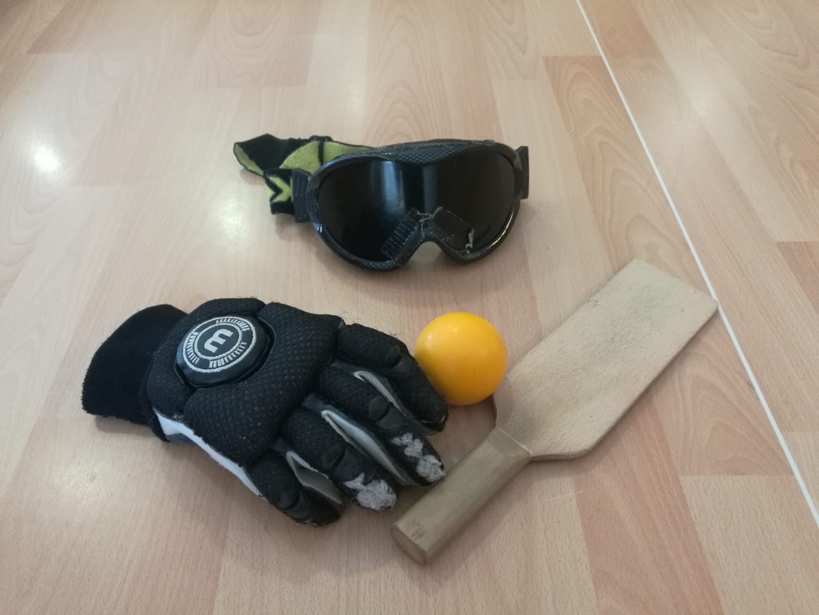 Se ve material de Showdown sobre un suelo de madera. Incluye gafas protectoras, un guante acolchado, una pala de madera y una pelota amarilla. Todo el conjunto está dispuesto junto, mostrando el equipamiento básico de este deporte.