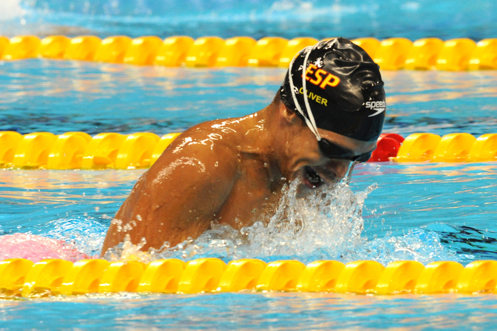 Una persona nada en una piscina realizando un estilo de braza, con gorro negro y gafas. El cuerpo aparece inclinado hacia adelante mientras el agua salpica alrededor del rostro y los hombros. Se ven corcheras amarillas delimitando el carril sobre el fondo azul del agua.