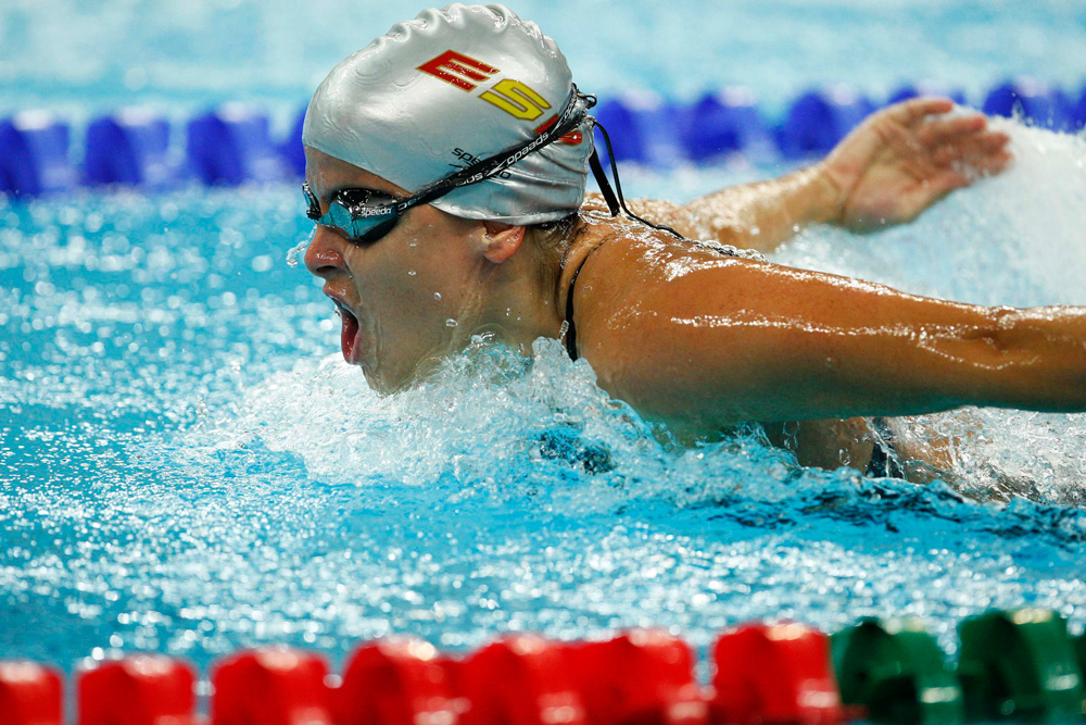 Una persona nada estilo mariposa en una piscina, con gafas y gorro. El agua salpica alrededor mientras respira y avanza por su carril. Se ven flotadores rojos y un fondo azul desenfocado.