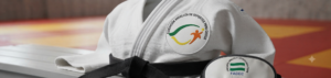 Un judogi blanco con cinturón negro descansa sobre un banco junto a un antifaz, ambos con el logotipo de la Federación Andaluza de Deportes para Ciegos (FADEC).
La imagen está tomada en un tatami de judo y simboliza la práctica deportiva adaptada para personas con discapacidad visual.